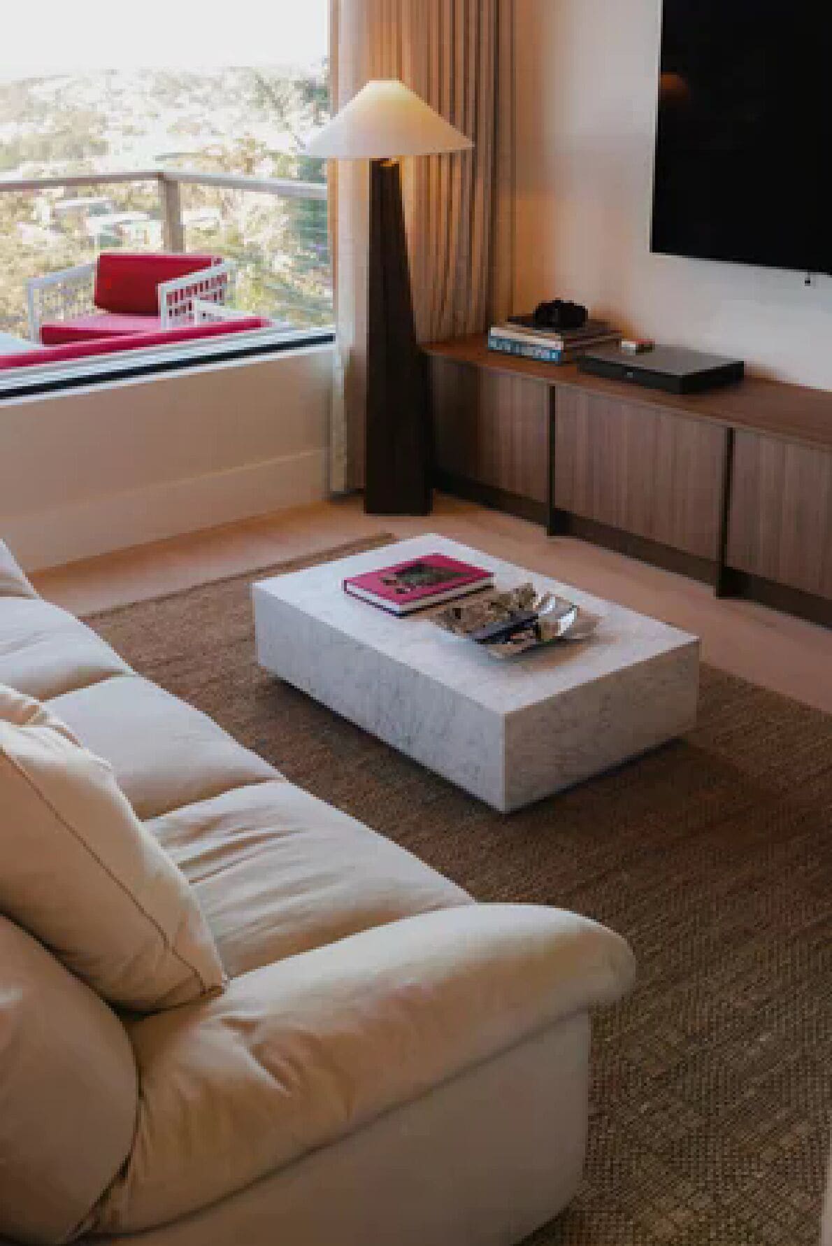 A modern living room with a sofa, a coffee table, a lamp, a flat-screen TV, and a view of the outdoors.