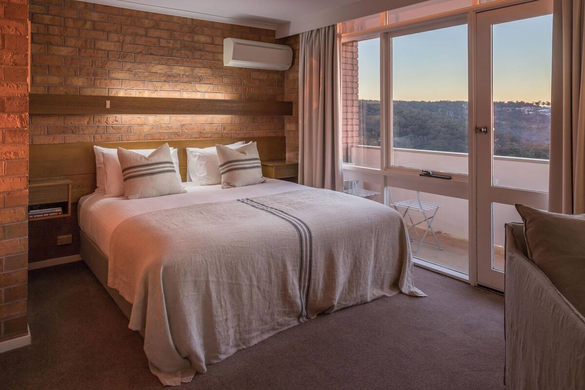 Une chambre avec un mur en briques, un lit avec une literie blanche, une fenêtre avec des rideaux et une vue sur l’extérieur.