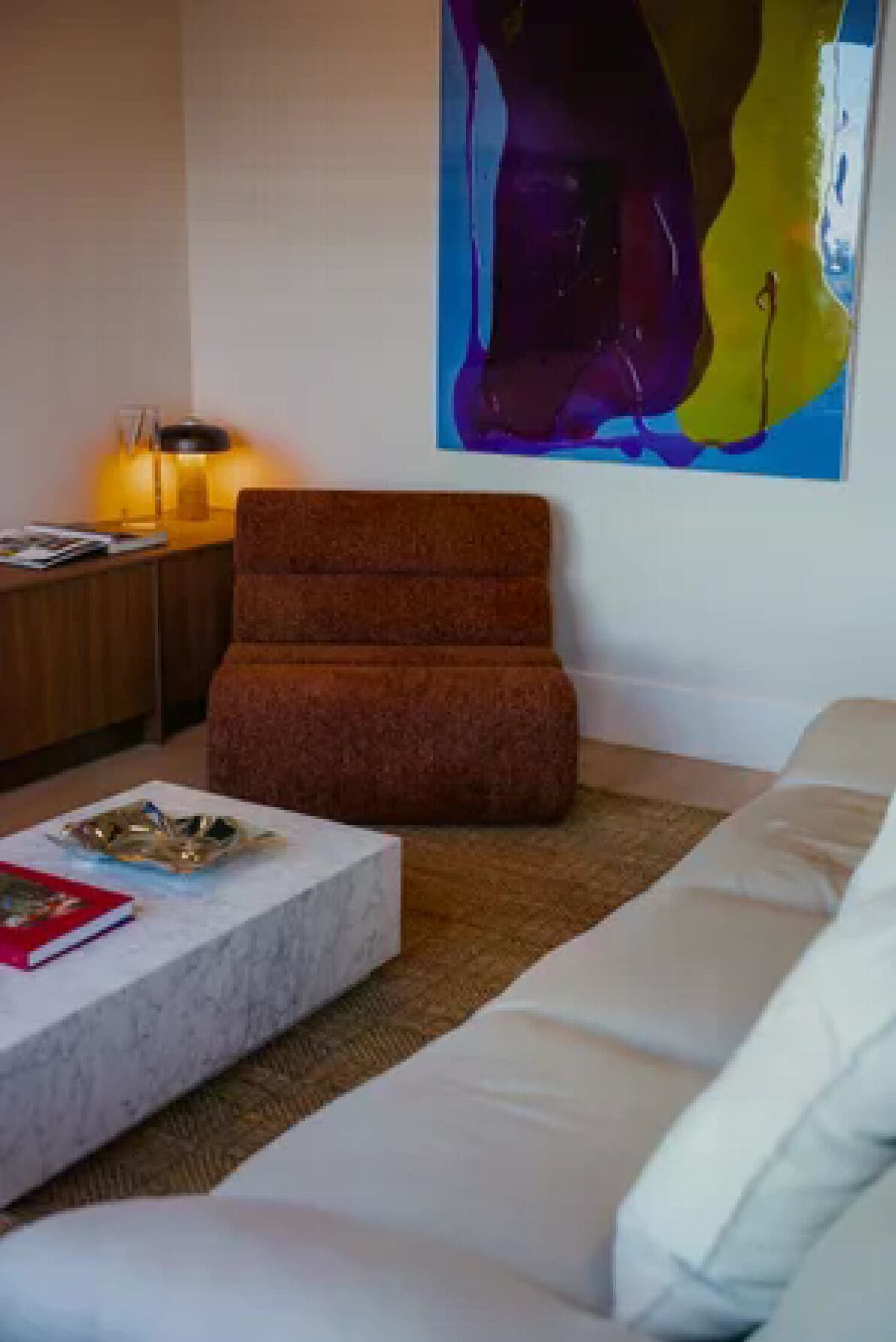 Un salon avec un canapé marron, un divan blanc, une table basse avec des magazines, une lampe et un grand tableau abstrait accroché au mur.