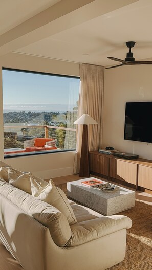 A modern living room with a large window, a flat-screen TV, a sofa, a coffee table, and a lamp. - Hillcrest Merimbula (Merimbula)