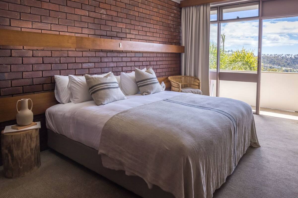 Une chambre avec un mur en briques, un grand lit avec une literie blanche, une tête de lit en bois, une chaise en osier et une vue dégagée sur l’extérieur grâce à une grande fenêtre.