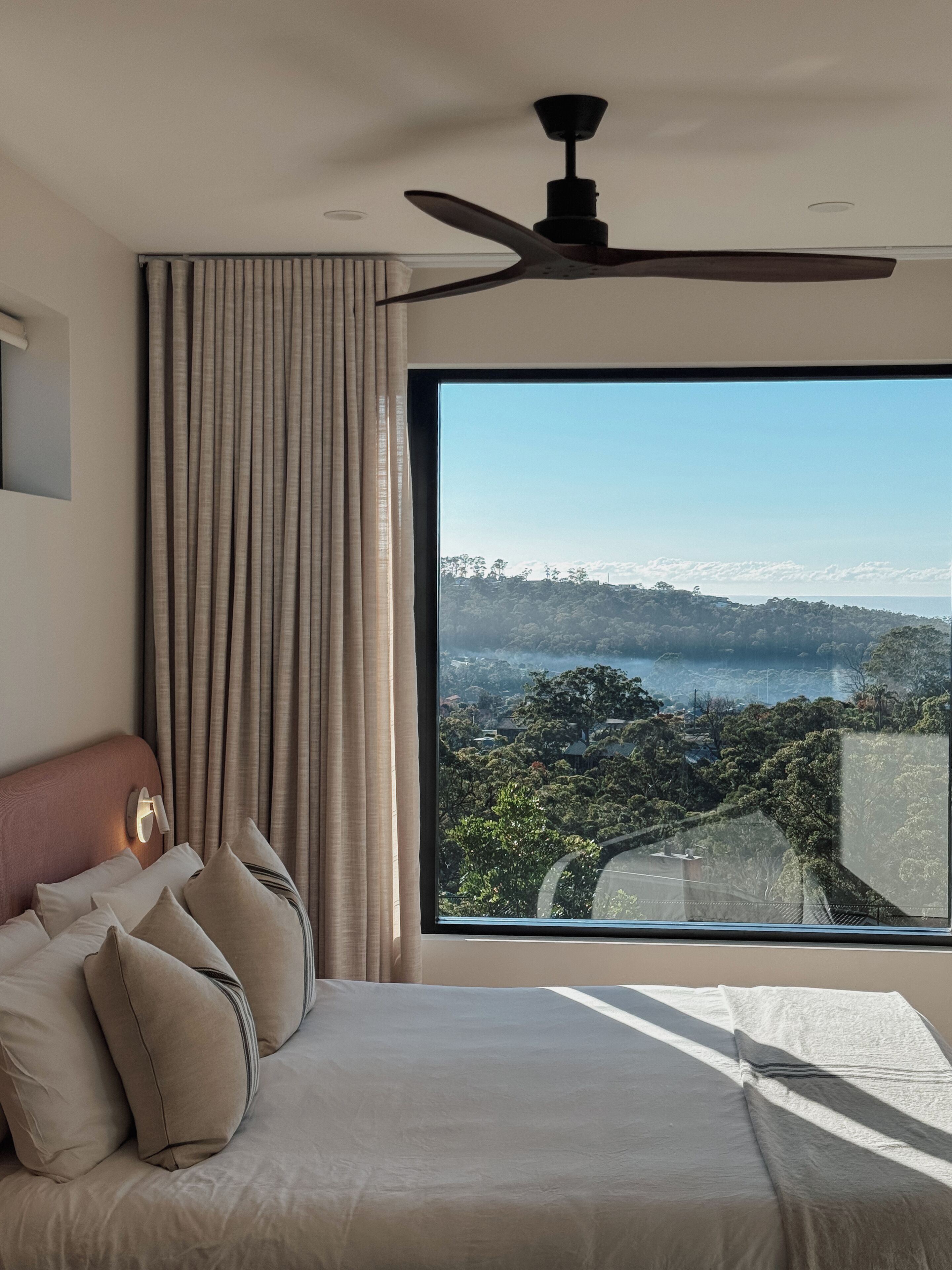 A bedroom with a large window, a ceiling fan, and a bed with pillows.