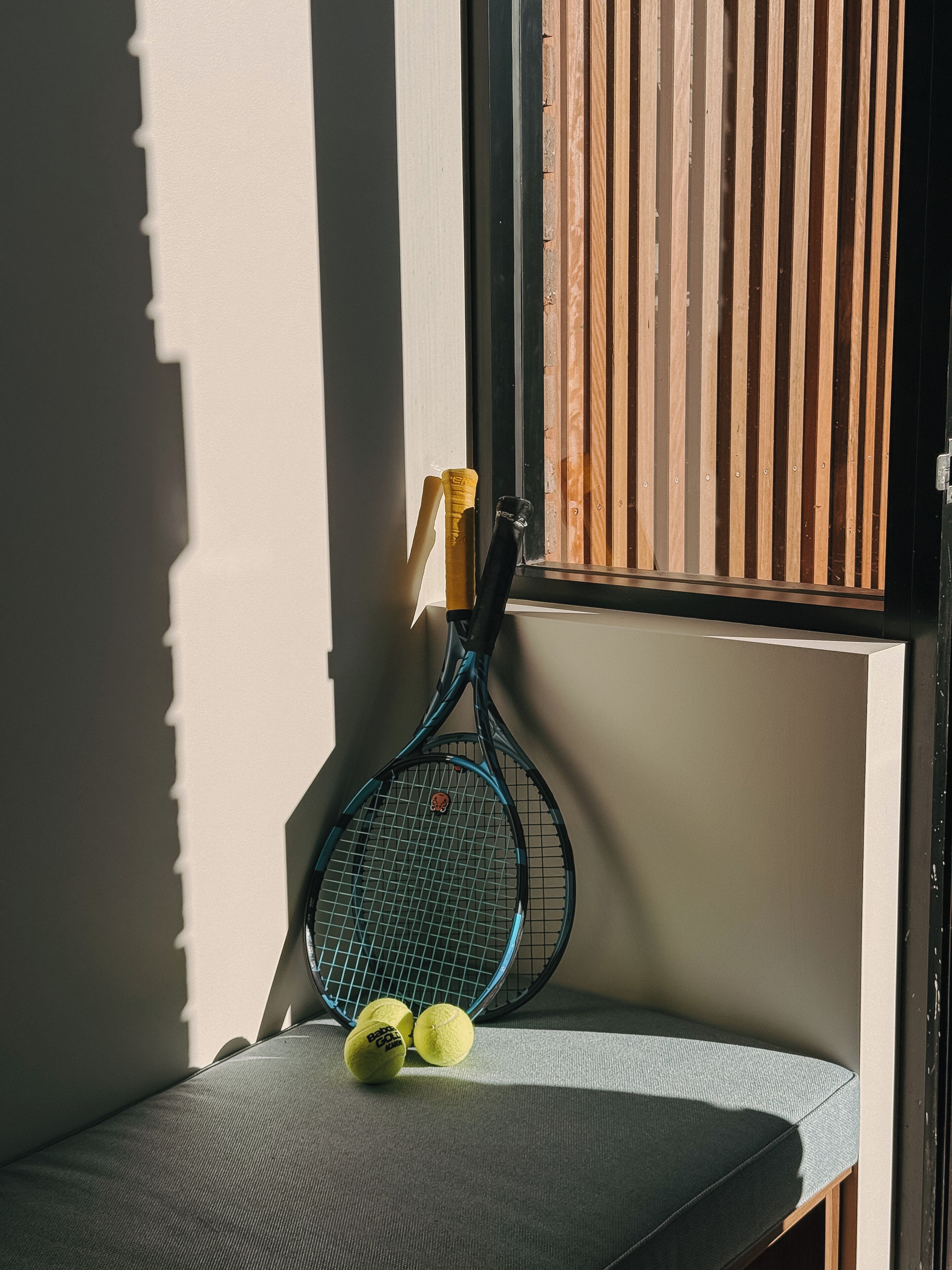 Una raqueta de tenis apoyada en una pared con una ventana, un cojín y dos pelotas de tenis sobre un banco.