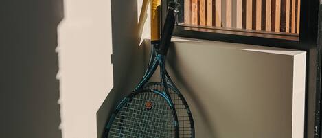 Una raqueta de tenis apoyada en una pared con una ventana, un cojín y dos pelotas de tenis sobre un banco.