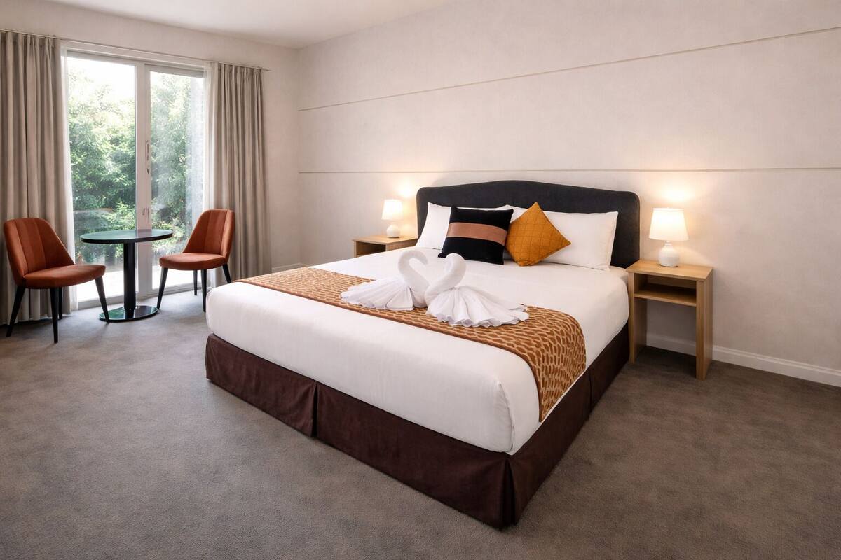 A hotel room with a large bed, two bedside tables with lamps, a small round table with two chairs, and a view of greenery through the window.