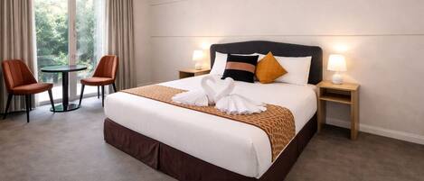 Habitación de hotel con una cama grande, dos mesitas de noche con lámparas, una mesita redonda pequeña con dos sillas y vistas a zonas verdes a través de la ventana.