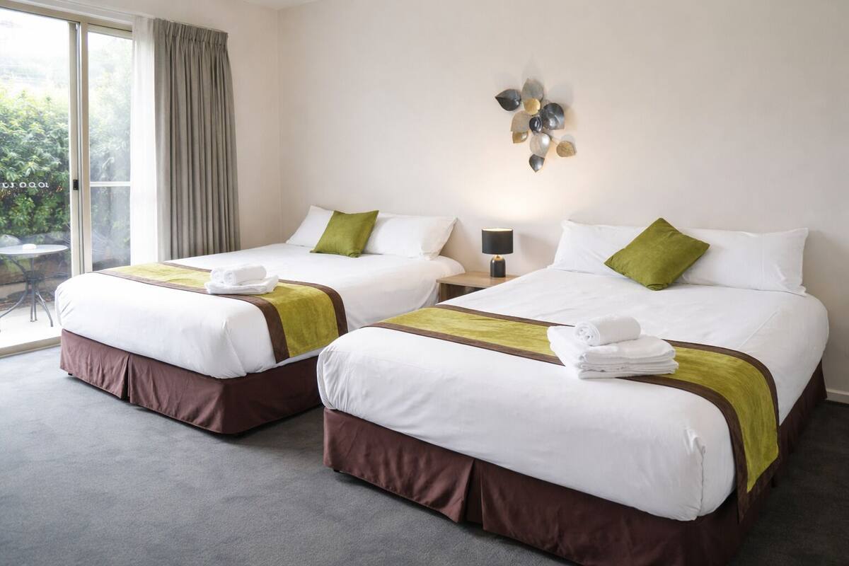 A hotel room with two beds, a small table, a lamp, and a view of greenery outside.