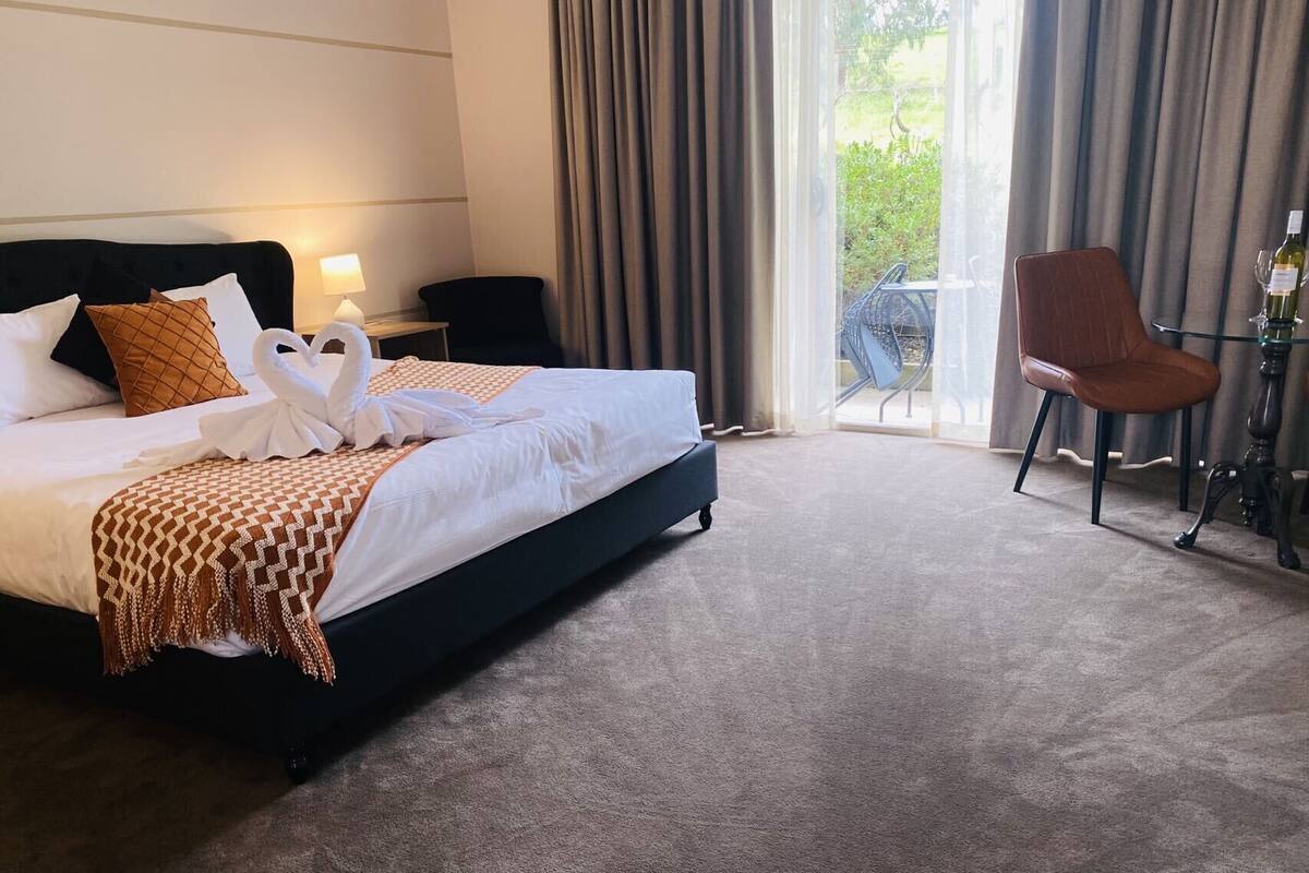 A hotel room with a bed, a chair, a table with a bottle, and a view of greenery outside.