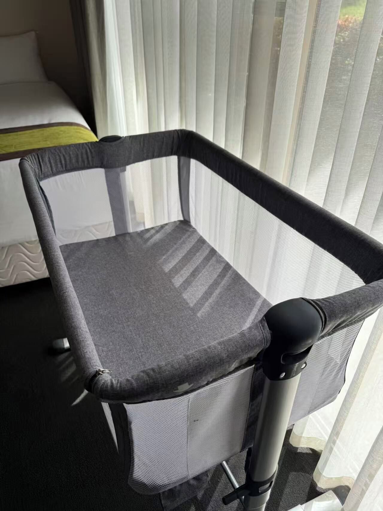A portable crib with a mesh base and a grey fabric top, positioned near a window with vertical blinds.