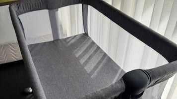 A portable crib with a mesh base and a grey fabric top, positioned near a window with vertical blinds.