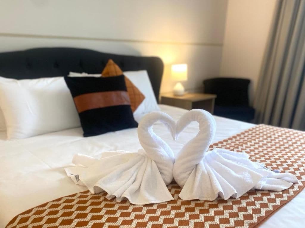 A hotel room with a bed, pillows, a nightstand, and a heart-shaped towel decoration.