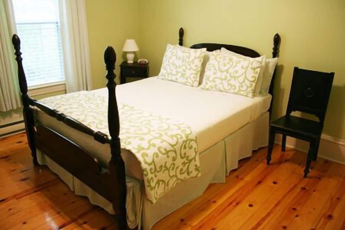 A neatly made bed with a wooden frame and a white bedspread with green patterns. There is a small bedside table with a lamp and a chair next to the bed.