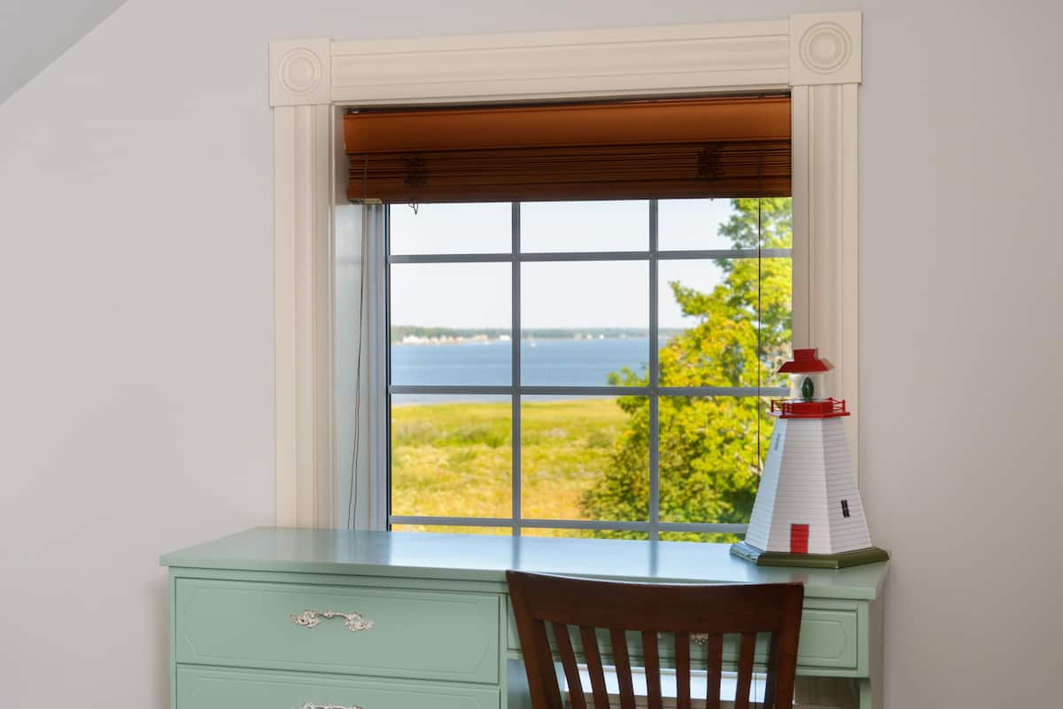 A desk with a view of the sea through a window with a white frame and brown blinds.