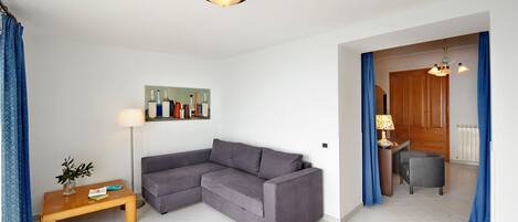 A modern living room with a grey sectional sofa, a wooden coffee table, and a floor lamp. There is a painting on the wall and a wooden door leading to another room.