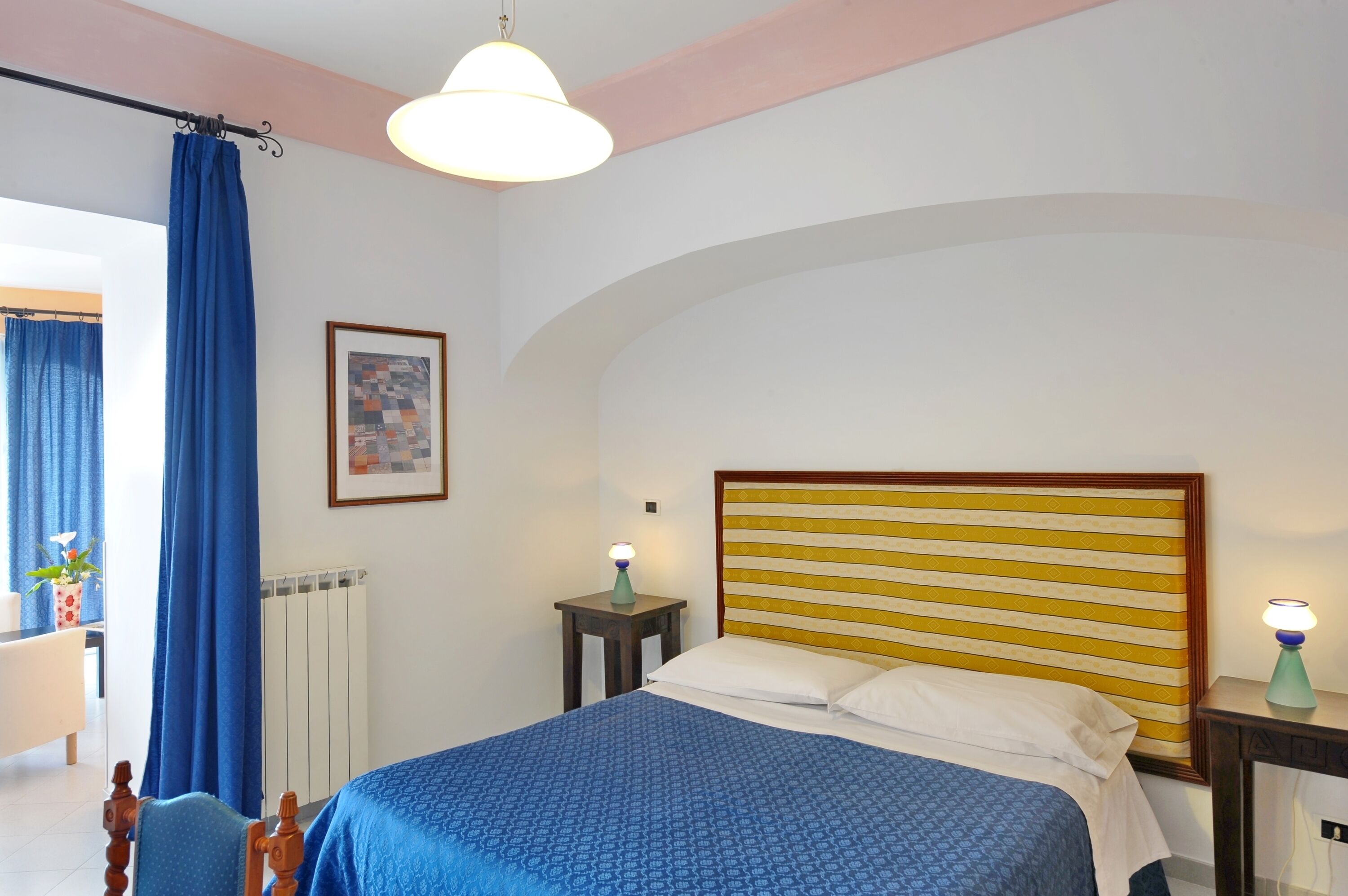 A bedroom with a bed, a nightstand, a lamp, a framed picture, and a window with curtains.