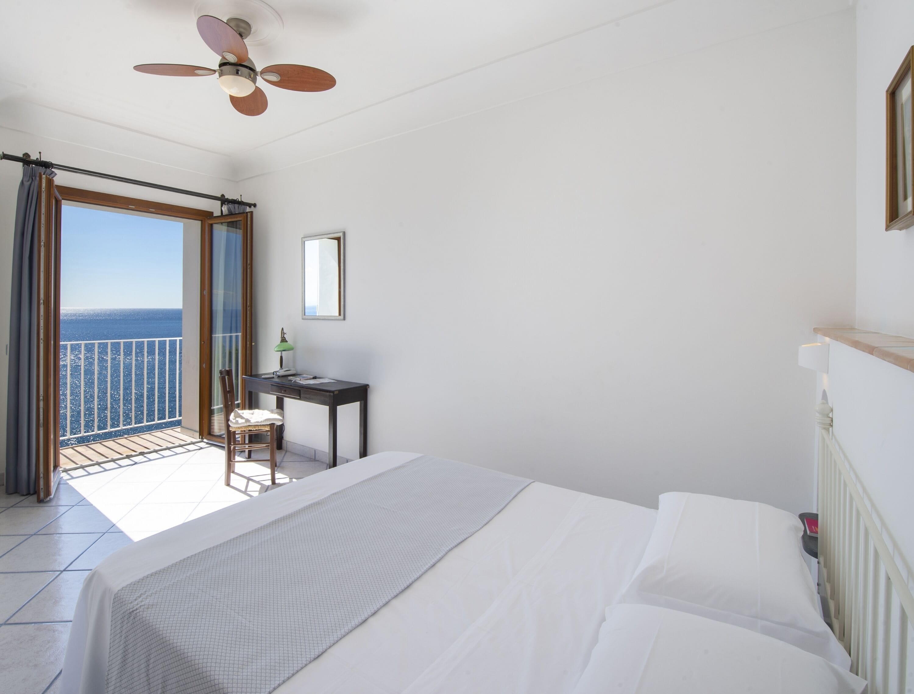 A bedroom with a bed, a desk, a chair, and a balcony with a view of the sea.