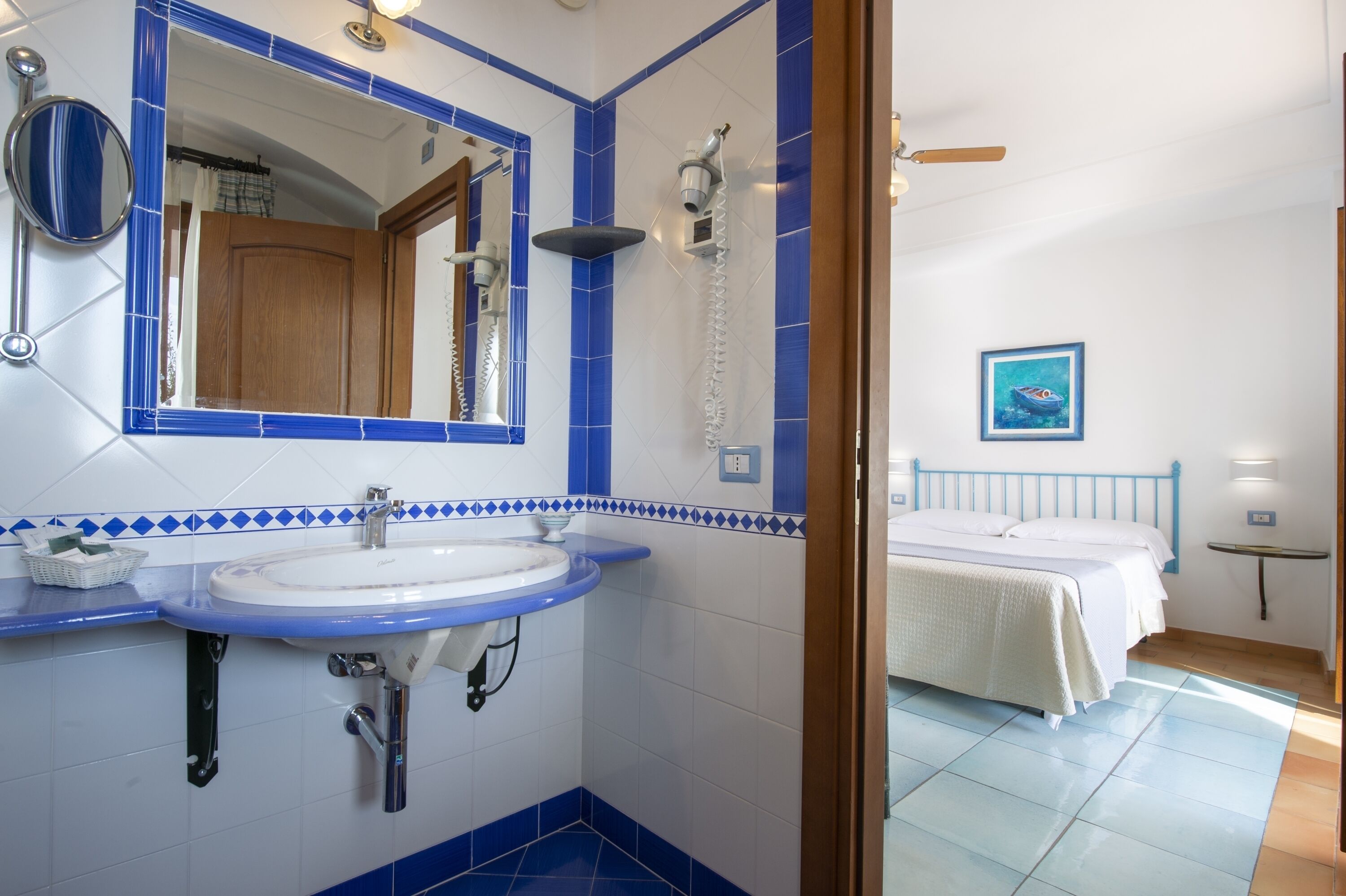 Un baño con lavamanos, espejo y ducha. Se ve una cama al fondo.