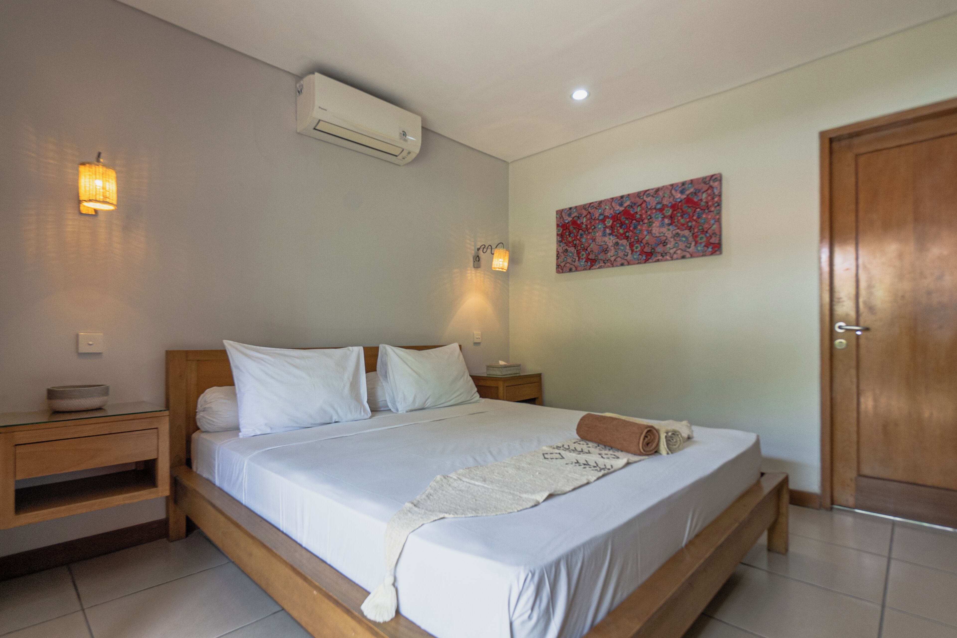 A bedroom with a wooden bed, white bedding, a wall-mounted air conditioner, two bedside lamps, a nightstand, and a door.