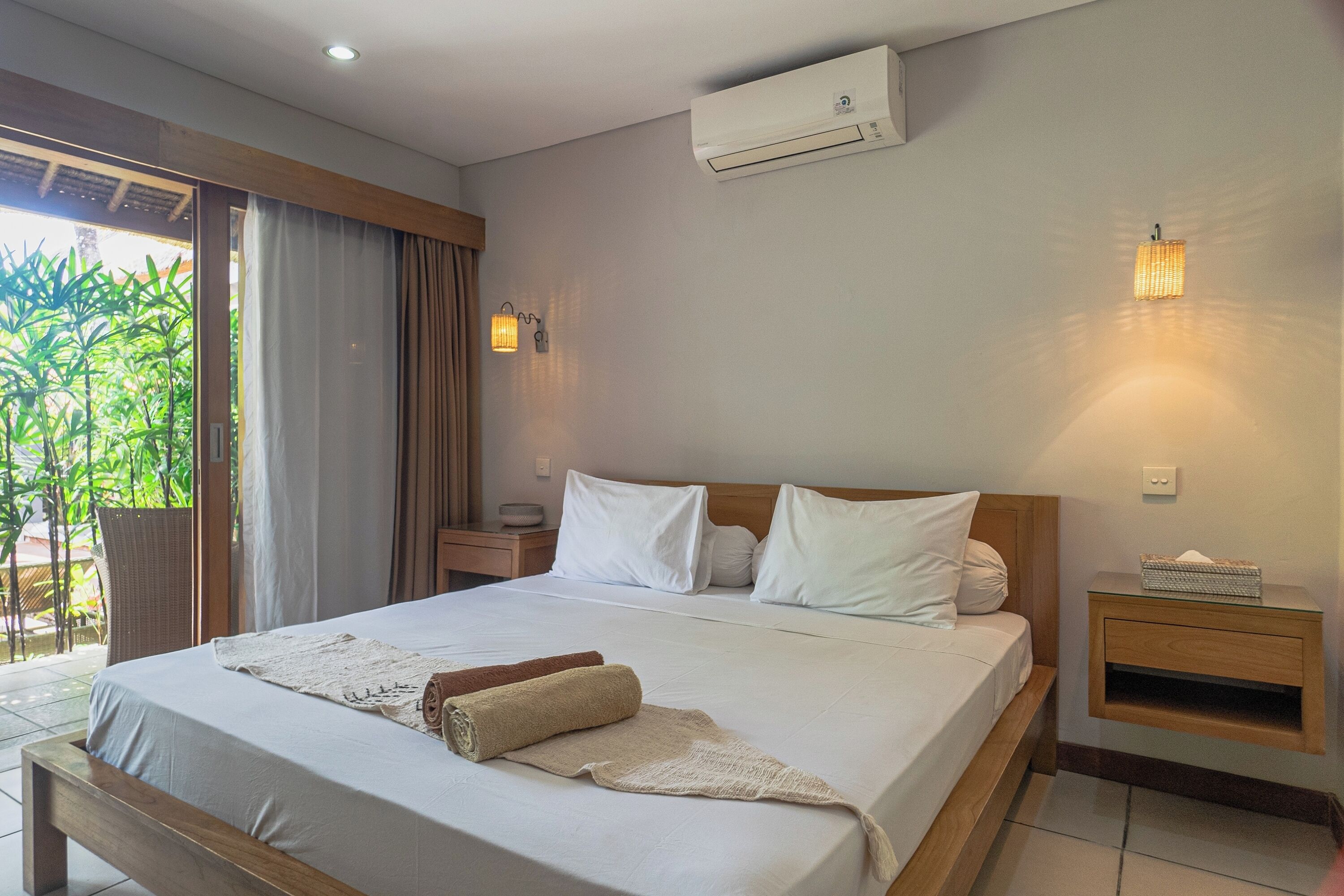 Una habitación de hotel con cama, mesita de noche, equipo de aire acondicionado y ventana con cortinas.