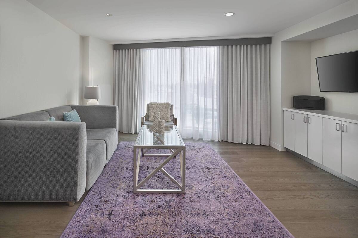 A modern living room with a grey sofa, a glass coffee table, a purple rug, a white cabinet, and a flat-screen TV.