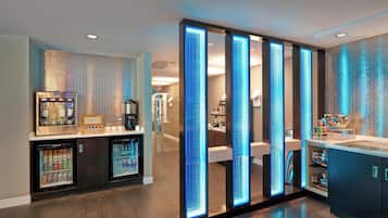 Un moderno vestíbulo de hotel con una estación de bebidas de autoservicio, vitrinas refrigeradas y un diseño elegante y contemporáneo.