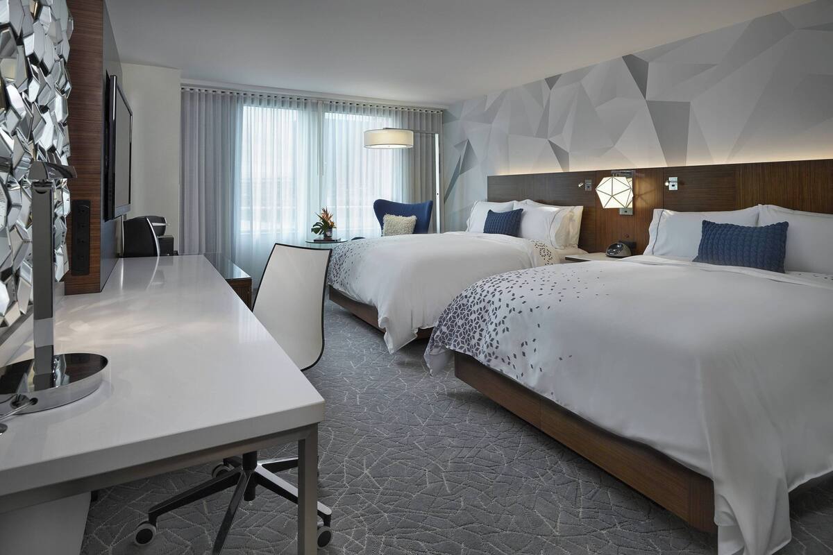 A hotel room with two beds, a desk, a TV, and a modern geometric wall design.