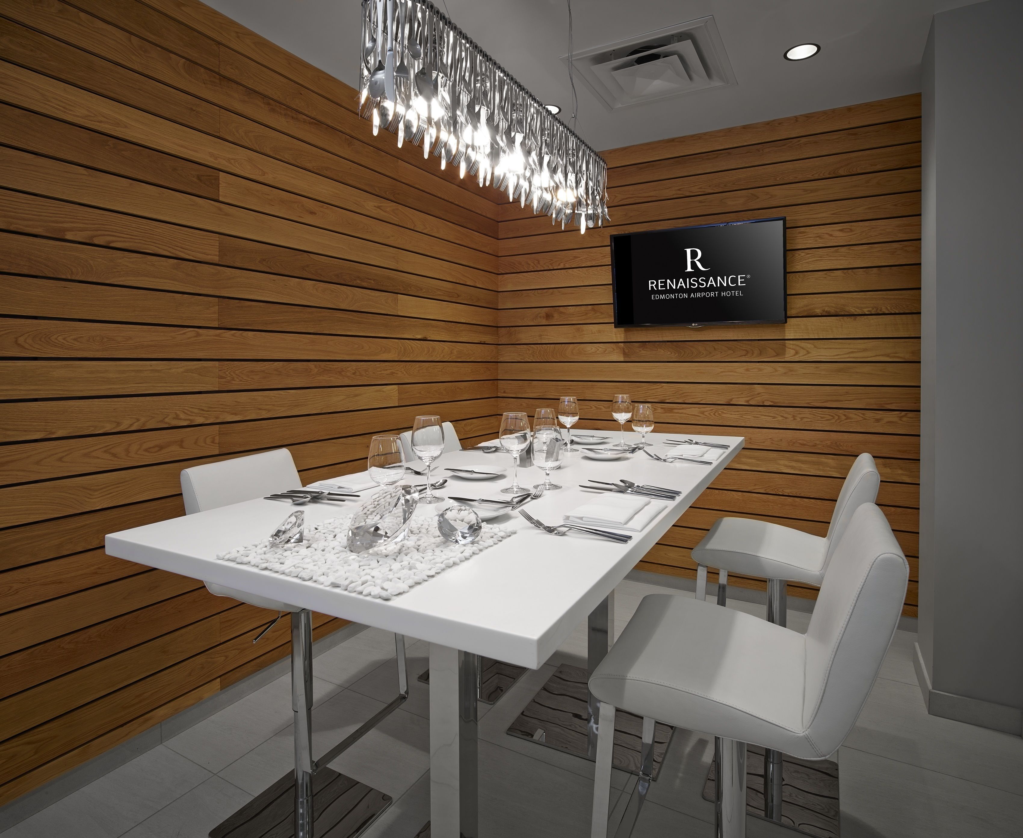 Un comedor moderno con una mesa blanca dispuesta para cuatro personas, rodeada de sillas blancas. Un candelabro cuelga encima, y un televisor en la pared muestra 