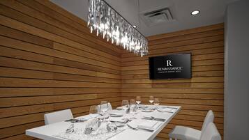 Un comedor moderno con una mesa blanca dispuesta para cuatro personas, rodeada de sillas blancas. Un candelabro cuelga encima, y un televisor en la pared muestra