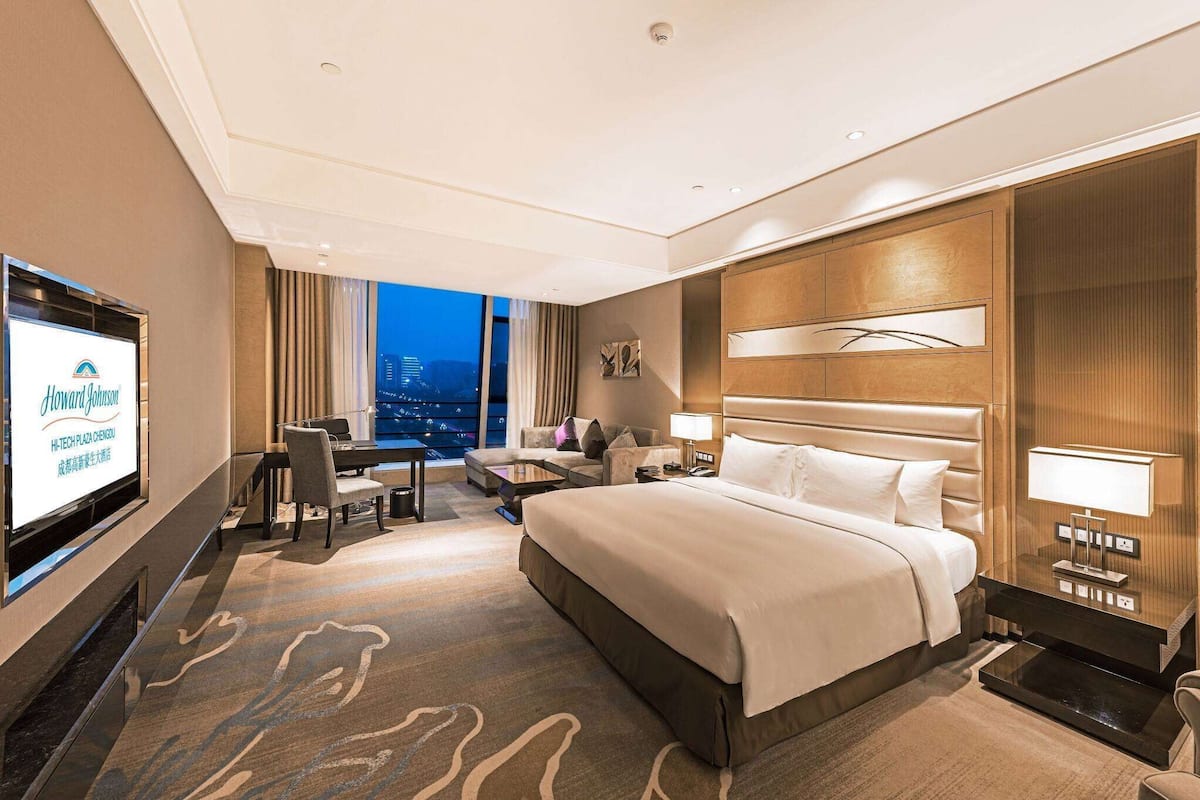 Ein Hotelzimmer mit einem großen Bett, einem Schreibtisch, einem Sessel, einem Fernseher und Blick auf die Stadt im Abenddämmerung.