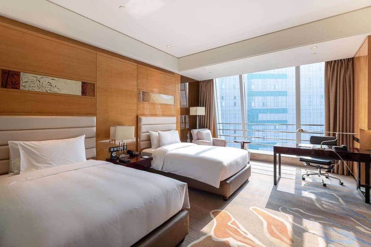 Ein Hotelzimmer mit zwei Betten, einem Schreibtisch, einem Stuhl und einem großen Fenster mit Blick auf die Stadt.