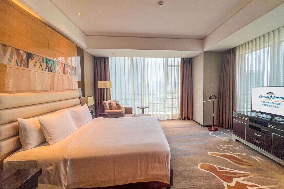 Ein Hotelzimmer mit einem großen Bett, einem Schreibtisch, einem Sessel, einem Fernseher und Blick auf die Stadt durch große Fenster.