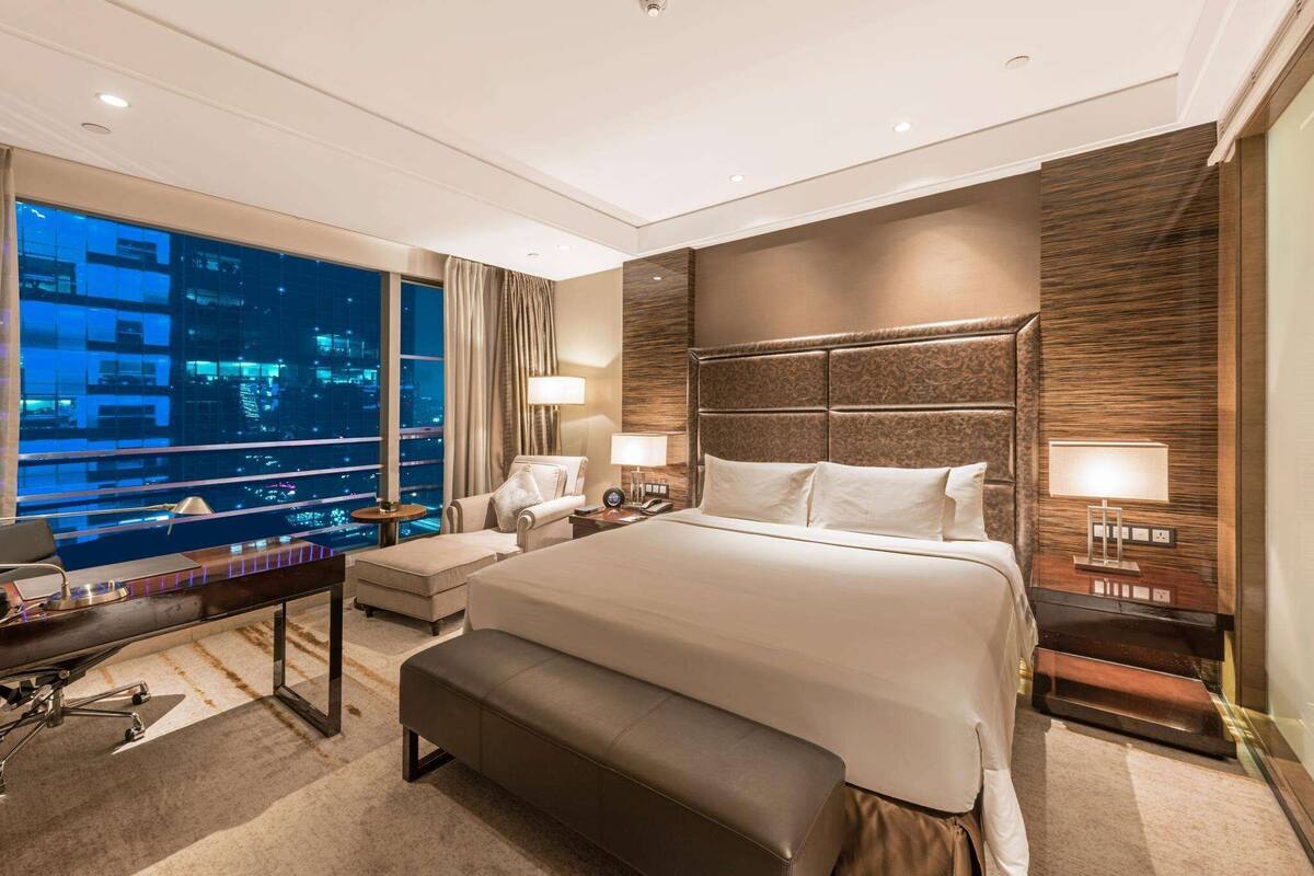 Ein modernes Hotelzimmer mit einem großen Bett, einem Schreibtisch, einem Stuhl und Blick auf die beleuchtete Stadt.