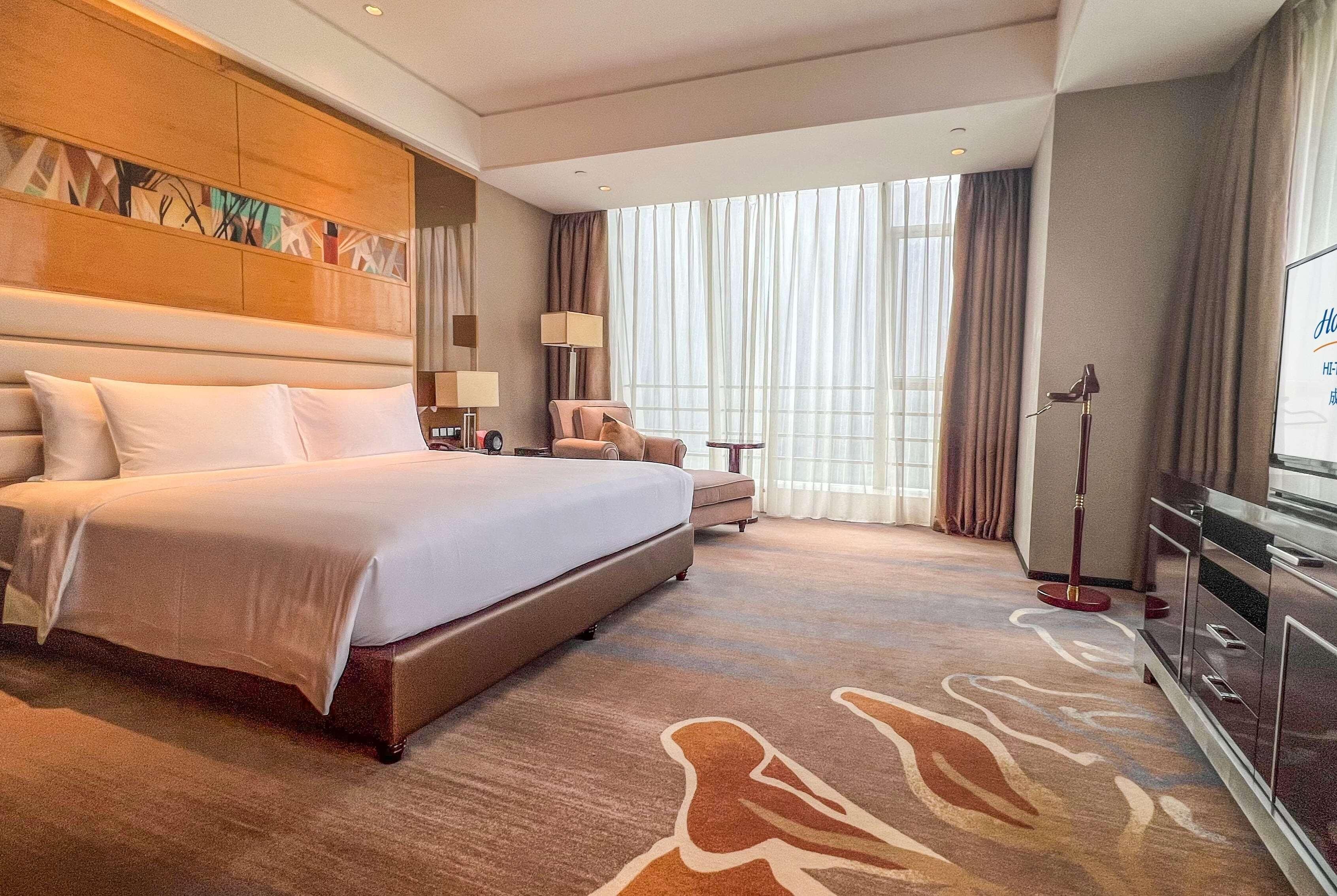 Habitación de hotel con una cama grande, un escritorio, una silla, un televisor y un ventanal con cortinas.