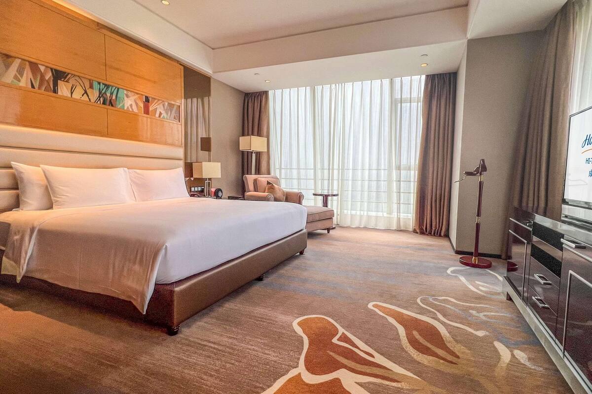 Habitación de hotel con una cama grande, un escritorio, una silla, un televisor y un ventanal con cortinas.