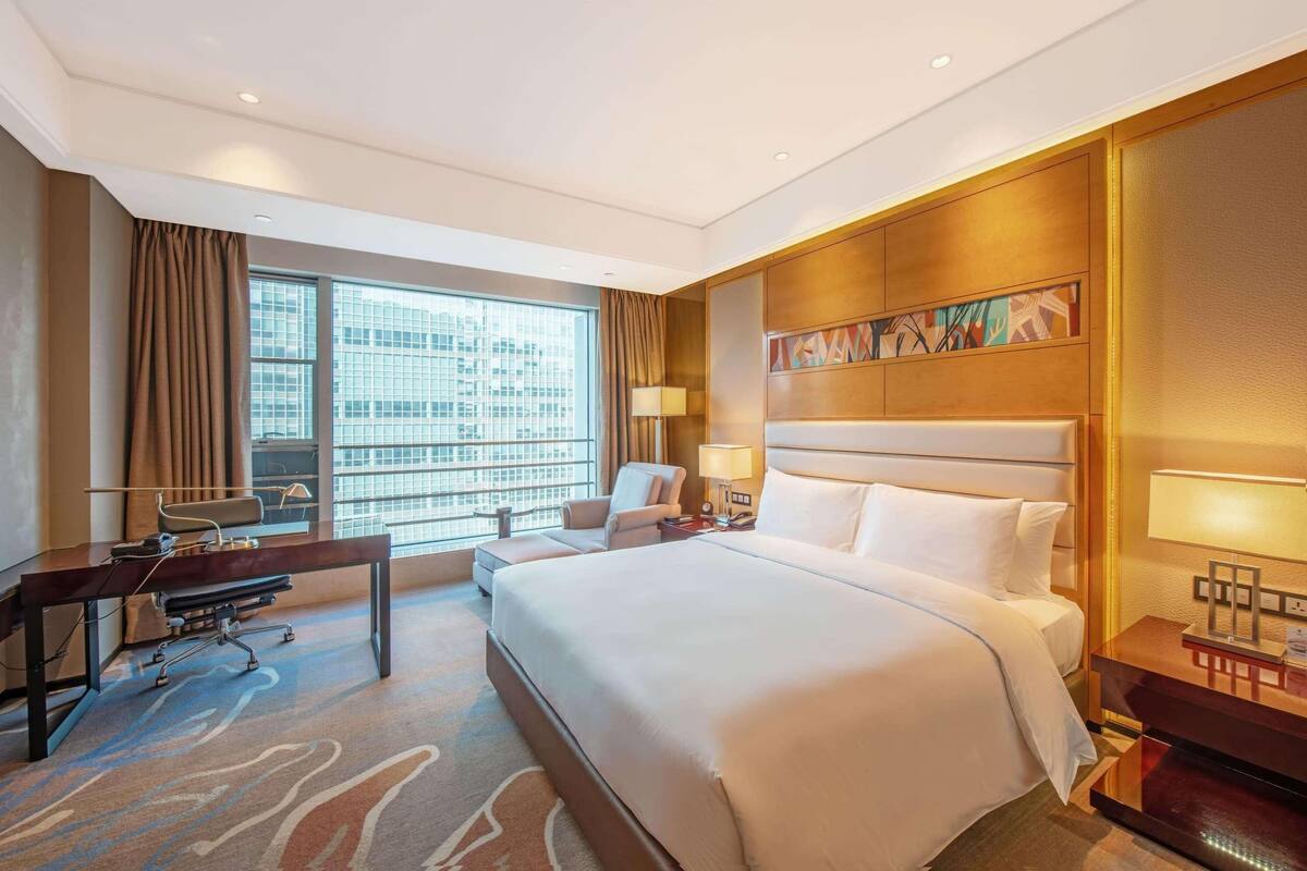 Habitación de hotel con una cama grande, un escritorio, una silla, una lámpara y vistas de la ciudad a través de amplios ventanales.