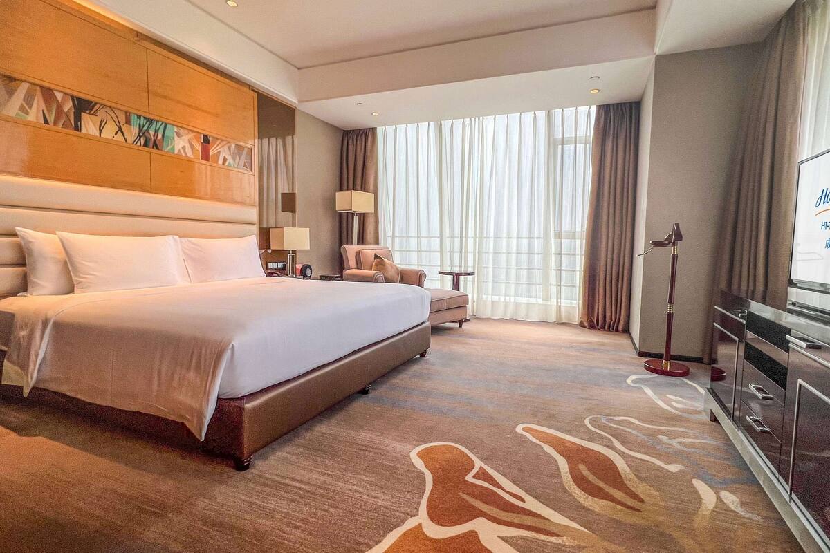 Ein Hotelzimmer mit einem großen Bett, einem Schreibtisch mit Stuhl, einem Fernseher und einem großen Fenster mit Vorhängen.