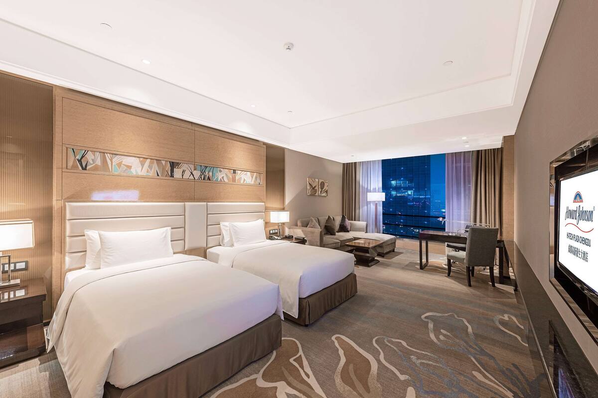Ein Hotelzimmer mit zwei Betten, einem Sofa, einem Fernseher und Blick auf die Stadt.