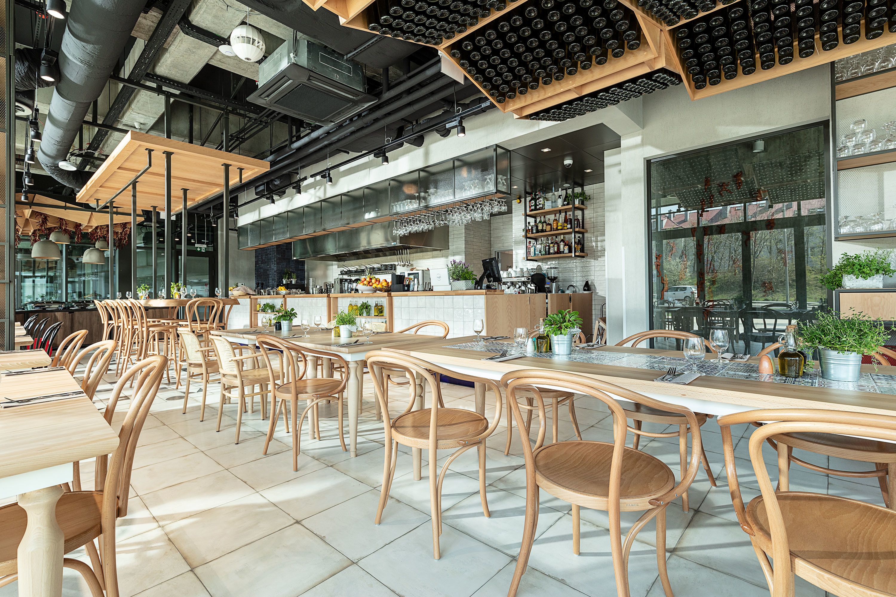 A spacious dining area with wooden tables and chairs, a kitchen in the background, and a modern industrial ceiling design.