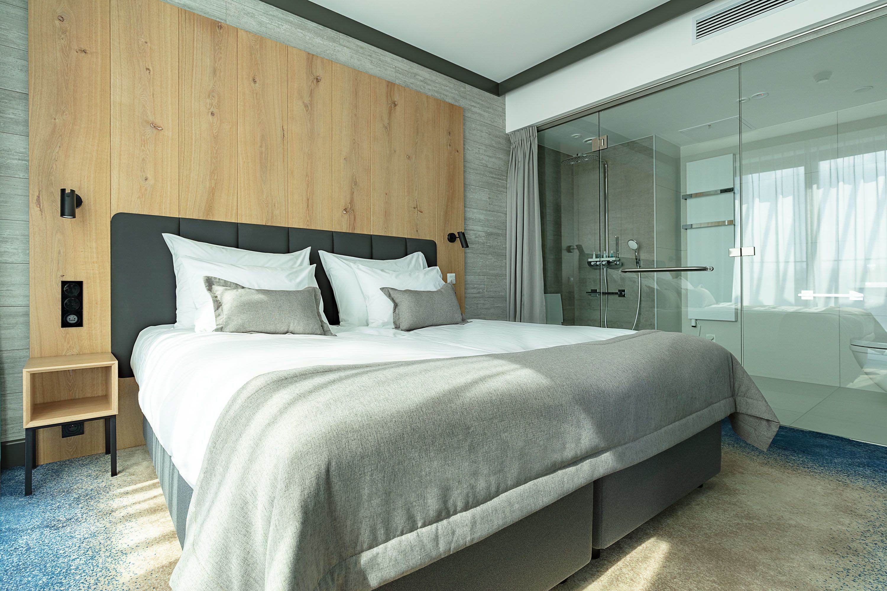 Ein modernes Hotelzimmer mit einem großen Bett, einer Wand mit Holzpaneelen, einer Glasdusche und einem kleinen Nachttisch.