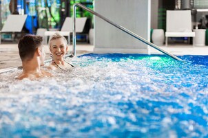 A couple in a hot tub with a metal railing, surrounded by lounge chairs and a pool area.