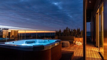 A hot tub on a wooden deck with a glass wall overlooking a forest and a sunset.