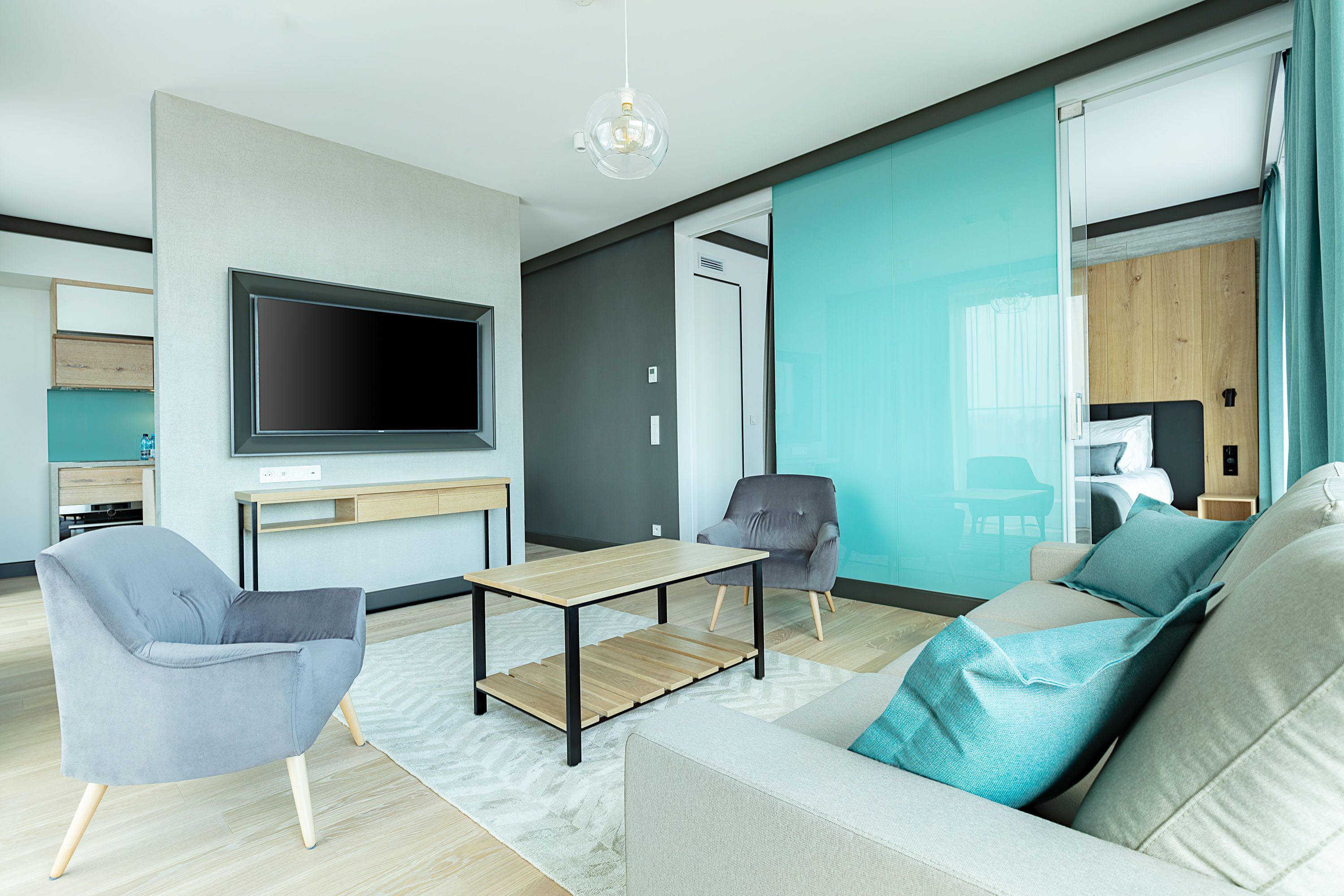 A modern living room with a flat-screen TV, grey armchairs, a wooden coffee table, and a sofa with teal cushions.