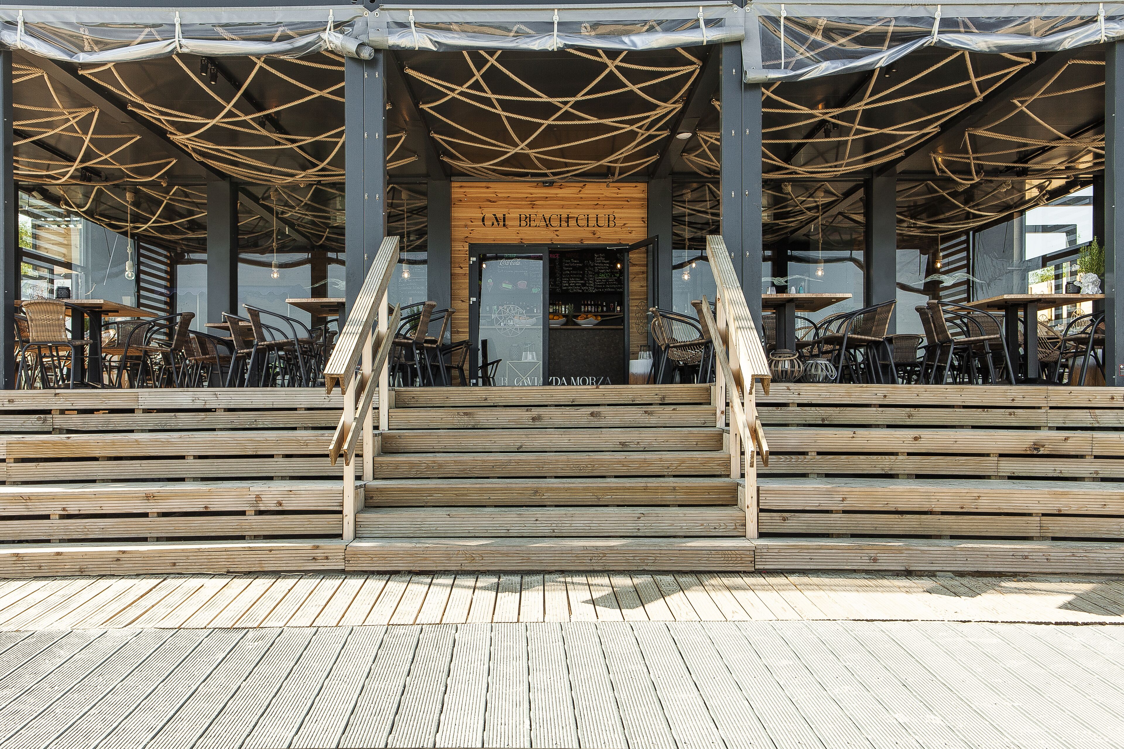Una terraza de madera con mobiliario exterior, una escalera de madera y un letrero que dice "GM BEACH CLUB".