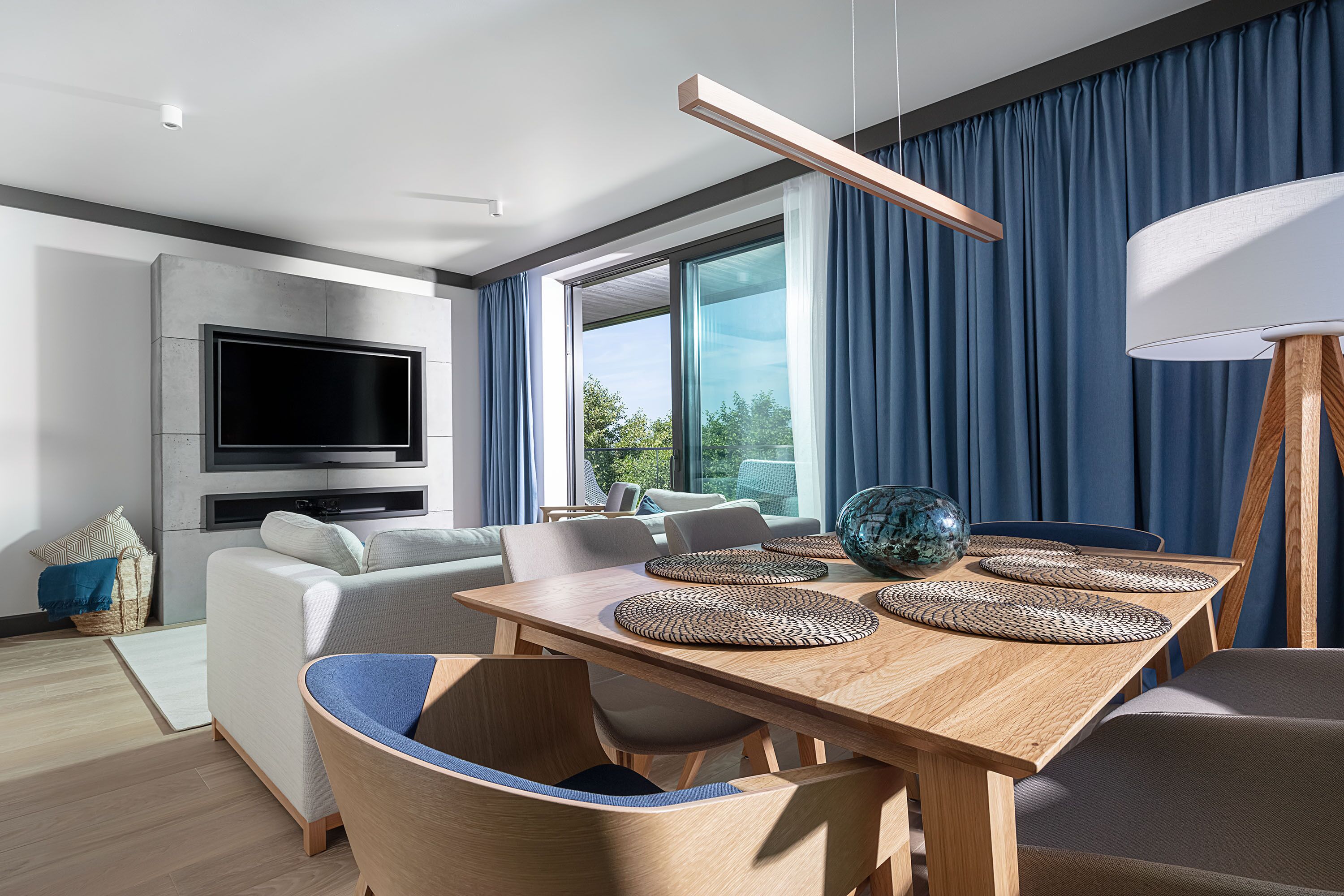 A modern living room with a flat-screen TV, a dining table with placemats, and a large window with curtains.