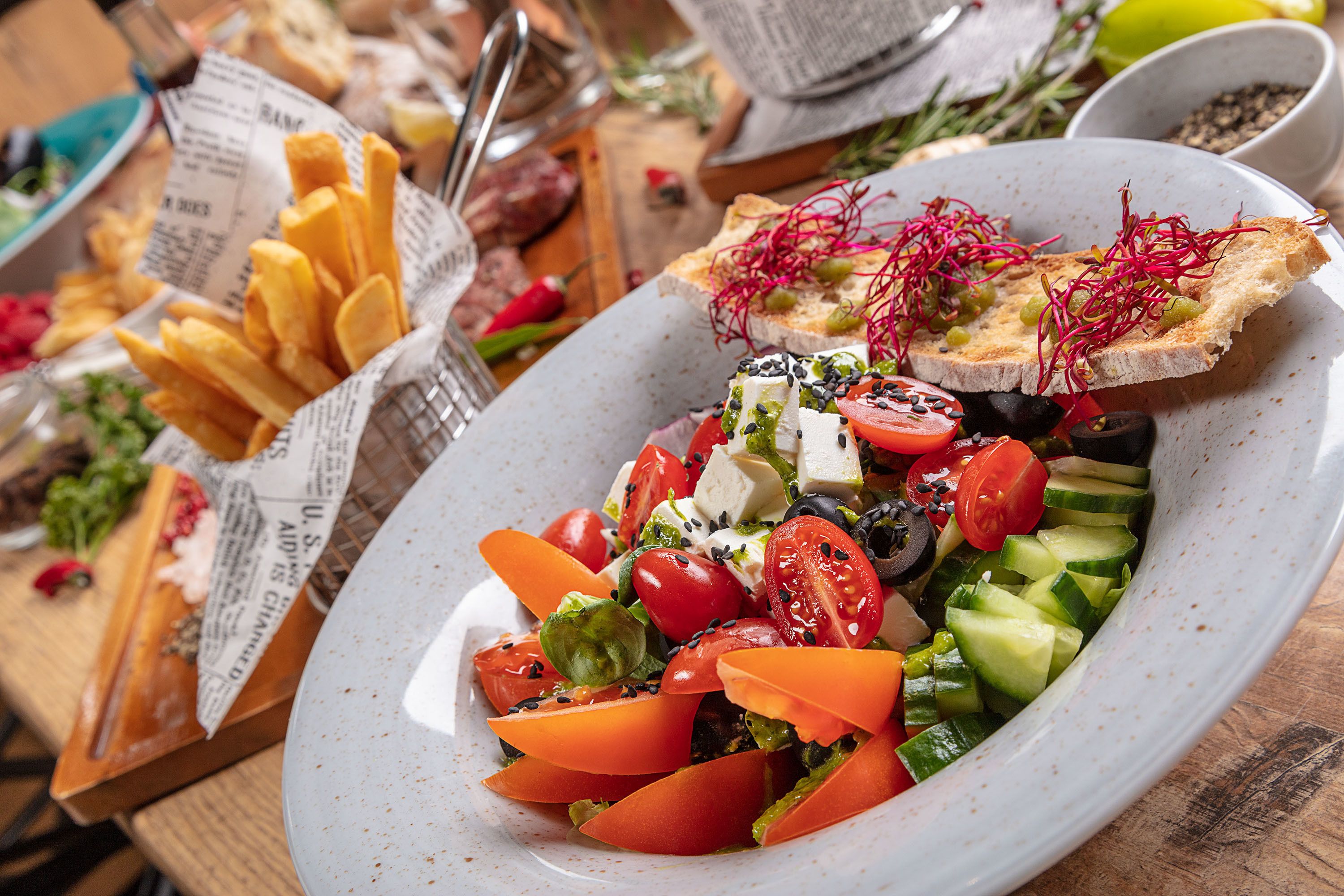 Une assiette de salade fraîche, composée de tomates, de concombres et de fromage, accompagnée d’une portion de frites et de tranches de pain garnies de germes.