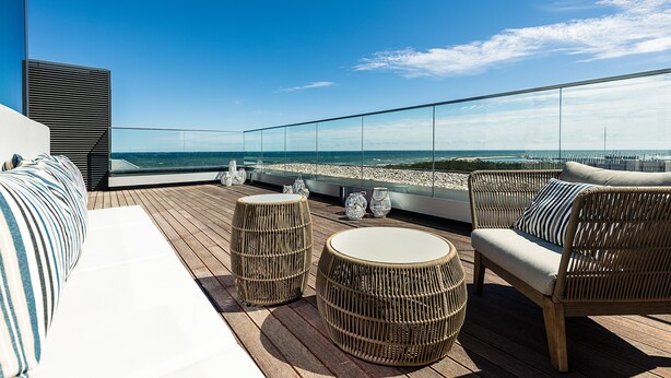 A rooftop terrace with wicker furniture, a glass balustrade, and a view of the sea.