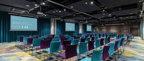 A conference room with rows of teal and purple chairs, a large screen, and a carpeted floor.