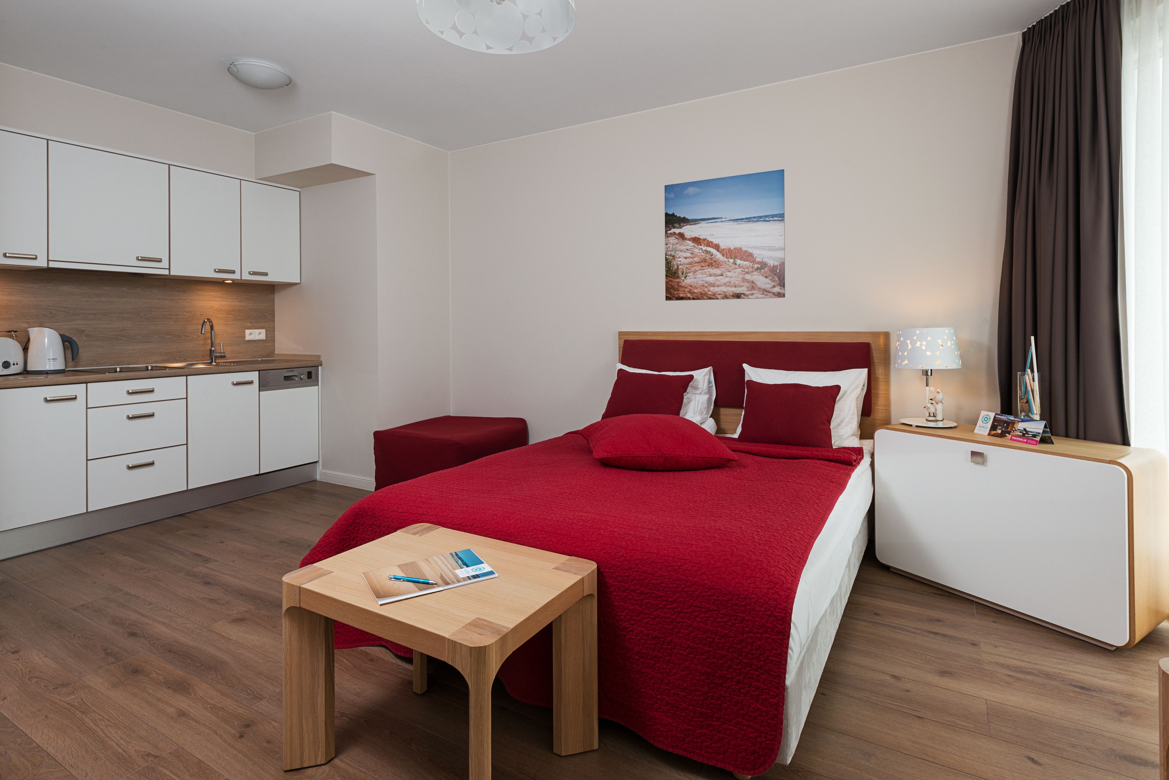 A modern hotel room with a wooden floor, a red bed with matching pillows, a small wooden table, a nightstand with a lamp, and a view of a snowy landscape through the window.
