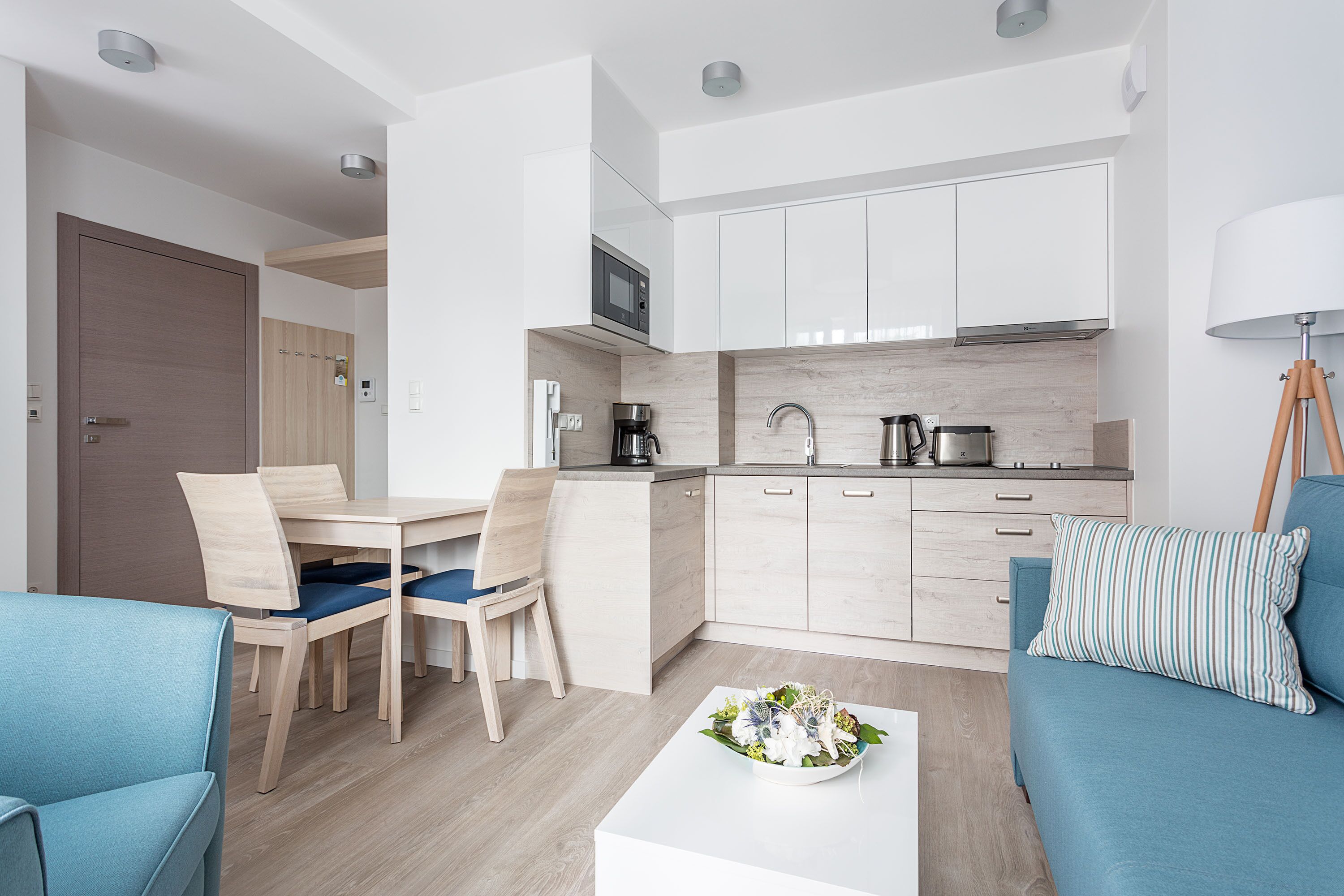 A modern kitchen with white cabinets, a microwave, and a coffee maker. There is a dining area with a table and chairs, and a blue sofa with a striped pillow.