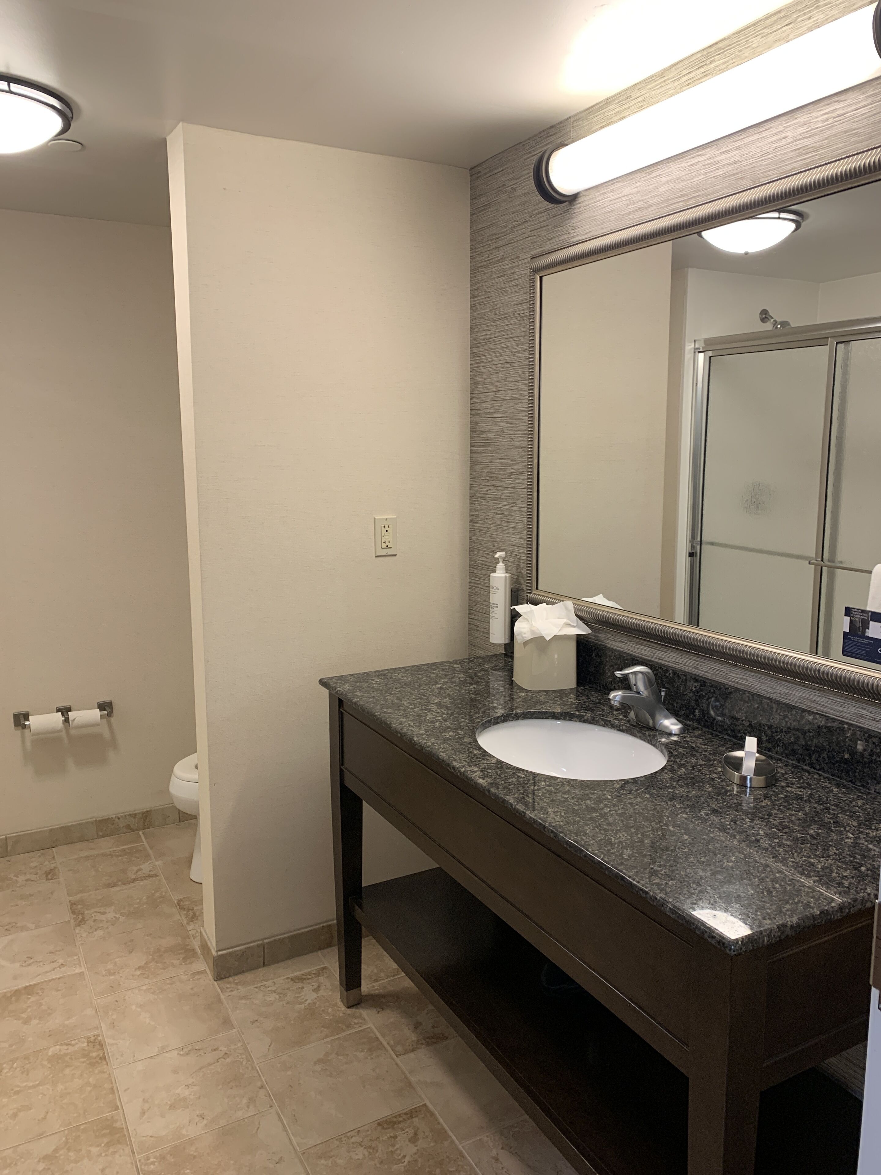 Un baño con lavamanos de encimera de granito, un espejo y una ducha a la vista al fondo.